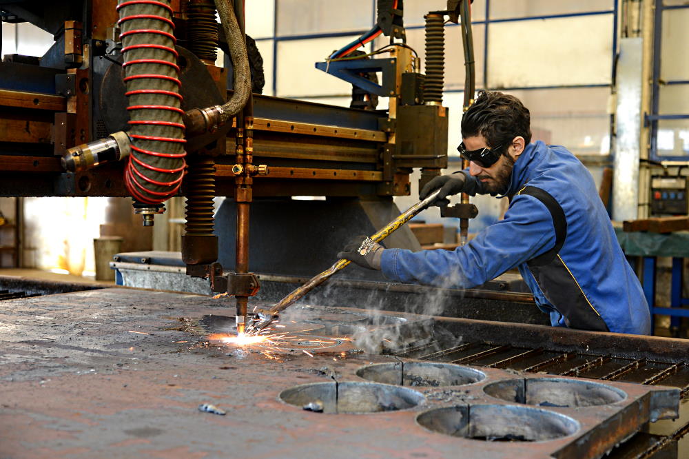 Un ouvrier soude une pièce métallique dans une usine. Il porte des lunettes de sécurité.