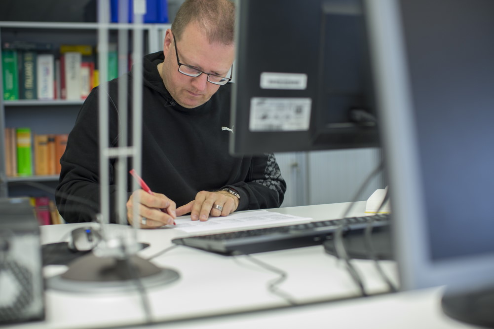 Un homme devant un ordinateur corrige un document imprimé avec un stylo rouge.