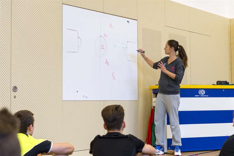 Dans une salle de gymnastique, une enseignante explique à ses élèves les règles d’un sport sur un tableau.