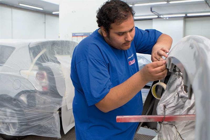 Un apprenti protège certaines parties d'une carrosserie avec du scotch de carrossier avant de la vernir.